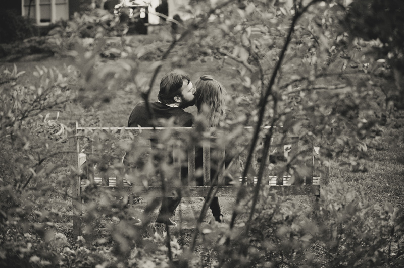 A black and white photograph of a couple kissing while sitting on a bench in Alumni Hall gardens at Penn State while their photographer photographs their engagement session.